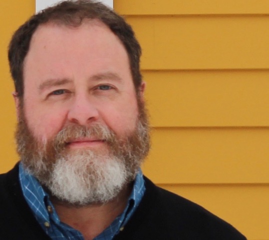headshot of a man with a graying beard in front of a mustard yellow house