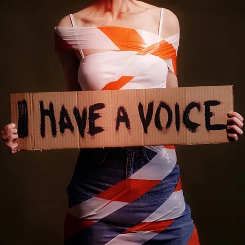 body of a woman wrapped with tap holding a cardboard sign saying "I Have a Voice"