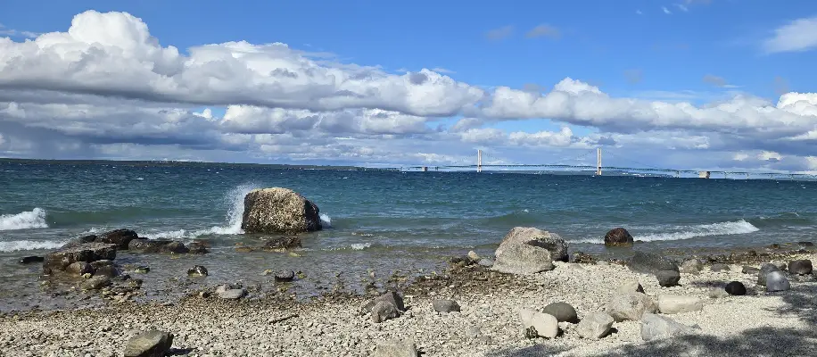 Rocky shoreline at McGulpin Point with Lake Michigan in the background and the Mackinac Bridge stretching across the horizon under a partly cloudy sky.