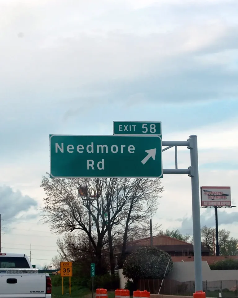 A green highway exit sign indicating "Exit 58 Needmore Rd" with an arrow pointing to the right. The sign is mounted on a metal pole, and there are trees and buildings in the background.